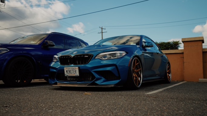 A blue sports car with customized details is parked next to a larger blue vehicle. The car has a personalized license plate and distinctive bronze-colored rims. The setting appears to be an outdoor parking lot with clear skies and scattered clouds.
