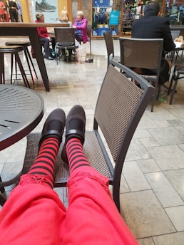 A view from someone's perspective sitting in a mall, wearing red pants and black shoes with red and black striped socks. In front, there is a seating area with three women chatting together at a table and a man facing away in a black jacket. The background displays a shoe store and some other shops.