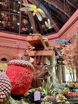 A vibrant indoor garden scene featuring giant acorns, colorful artificial dragonflies, a waterfall, and lush greenery. The setting is under a glass-domed ceiling with intricate metal framework. Various plants and decorative elements add to the ambiance, alongside a sign requesting visitors not to walk through the garden.