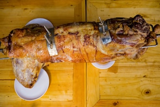 A whole roasted pig on a rotisserie is placed on a wooden table. The skin is golden and crispy, and two metal brackets are securing it in place on the spit. The setting suggests a rustic or outdoor environment.