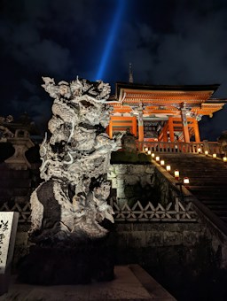 An intricately carved dragon statue stands prominently in front of an illuminated traditional Asian temple. The structure is accentuated with vibrant orange and wooden textures. A flight of stone steps leads up to the temple, lined with glowing lanterns that provide a warm, inviting glow. The night sky is partially clouded, with a bright beam of light projecting upwards, creating a mystical atmosphere.