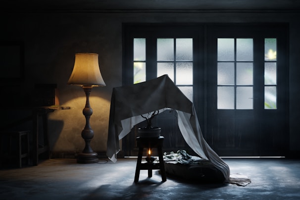 A dimly lit room featuring a tall floor lamp with a warm glow, casting soft shadows on the walls. A small potted plant rests atop a stool, partially covered by a draped white cloth. In the background, large frosted glass doors allow some natural light to enter, creating a gentle contrast between the warm interior and the cool exterior lighting.