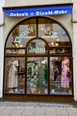 A storefront with an arched glass window displays traditional dresses on mannequins. The window showcases three dirndls in white, green, and pink, along with decorative items and a sign indicating '60 Jahre'. Above the window, a blue awning bears the words 'Indra's Dirndl-Ecke' in white lettering.