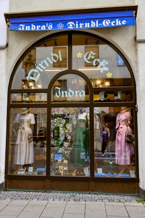 A storefront with an arched glass window displays traditional dresses on mannequins. The window showcases three dirndls in white, green, and pink, along with decorative items and a sign indicating '60 Jahre'. Above the window, a blue awning bears the words 'Indra's Dirndl-Ecke' in white lettering.