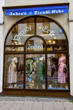 A storefront with an arched glass window displays traditional dresses on mannequins. The window showcases three dirndls in white, green, and pink, along with decorative items and a sign indicating '60 Jahre'. Above the window, a blue awning bears the words 'Indra's Dirndl-Ecke' in white lettering.