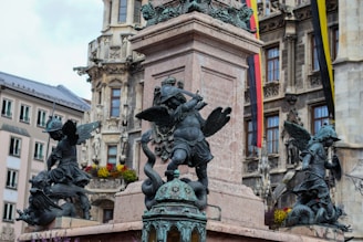 The image features a detailed stone fountain adorned with several bronze statues of winged figures in a historical European setting. The figures appear to be engaged in battle with creatures or demons. Behind the fountain, a traditional building with ornate windows and balconies, along with colorful flags, enhances the scene.
