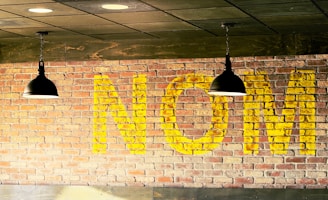 Large yellow letters spelling 'NOM' are painted on a brick wall. Two black pendant lights hang from the ceiling in front of the wall, with a high contrast between the dark fixtures and the warm, light tones of the brick and paint.