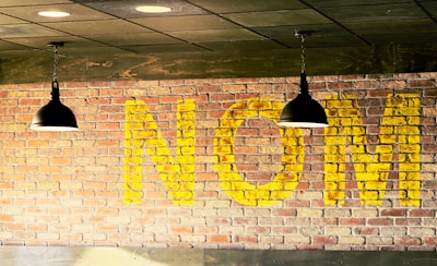 Large yellow letters spelling 'NOM' are painted on a brick wall. Two black pendant lights hang from the ceiling in front of the wall, with a high contrast between the dark fixtures and the warm, light tones of the brick and paint.