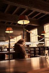 A cozy library setting with warm lighting from hanging lamps that have large shades. A giant teddy bear sits on a wooden table among rows of bookshelves filled with books. Colorful paper hearts are strung across the room, adding a playful atmosphere.