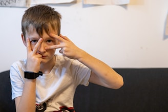 A young person wearing a white t-shirt sits on a dark couch. They are holding up both hands in front of their face, with their fingers forming V shapes around their eyes. The background shows a plain wall with some paper and drawings attached to it. The individual is wearing a black wristband or smartwatch on their left wrist.