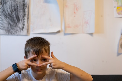 A child is making a gesture with both hands in front of their face while standing against a wall displaying several pieces of crayon and pencil artwork. The child wears a smartwatch on their wrist and is dressed in a white shirt.