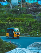 An auto rickshaw is navigating a curved road surrounded by lush greenery. The bright yellow vehicle contrasts against the dense vegetation and the clear blue sky above. The scene depicts a rural or suburban setting with a hint of infrastructure visible in the background.