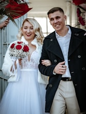 A couple smiling and walking arm in arm. The woman is holding a bouquet of red and white flowers and is wearing a wedding dress with lace details and a white fur wrap. The man is dressed in a white shirt and a dark coat.