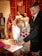 A woman in a white dress is signing a document on a table while holding a bouquet of red and white flowers. Several people stand around her, including a person pointing at the document. The room has wooden paneling and a warm ambiance.