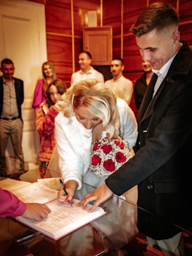 A woman in a white dress is signing a document on a table while holding a bouquet of red and white flowers. Several people stand around her, including a person pointing at the document. The room has wooden paneling and a warm ambiance.