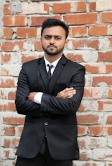 A man with a beard stands confidently with arms crossed in front of a textured brick wall. He is wearing a black suit and tie, with a white shirt peeking from underneath the suit jacket.