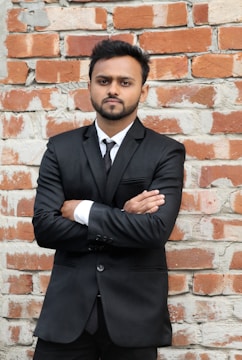 A man with a beard stands confidently with arms crossed in front of a textured brick wall. He is wearing a black suit and tie, with a white shirt peeking from underneath the suit jacket.