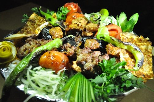 A vibrant and colorful platter containing a variety of grilled meats, vegetables, and garnishes arranged beautifully. The dish features succulent pieces of grilled meat, charred tomatoes, and peppers with sliced cucumbers and fresh herbs. Additional garnishes like lemon slices carved into flowers and bread pieces add to the presentation.