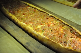 A long, rectangular flatbread topped with minced meat, diced vegetables, and herbs. It is placed on a wooden serving board and appears freshly baked, with a golden-brown crust.
