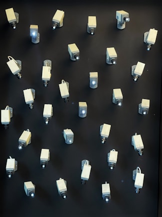 A wall is covered with multiple surveillance cameras arranged in a grid pattern. The cameras are uniform in shape and color, creating a stark and somewhat ominous appearance against the dark background.