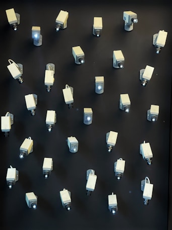 A wall is covered with multiple surveillance cameras arranged in a grid pattern. The cameras are uniform in shape and color, creating a stark and somewhat ominous appearance against the dark background.