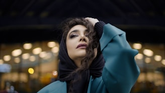 A person with a contemplative expression is wearing a teal overcoat and a black headscarf. Their hand rests on their head, and they stand against a backdrop of defocused lights, giving a bokeh effect.