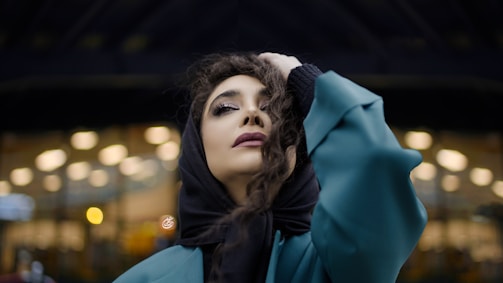 A person with a contemplative expression is wearing a teal overcoat and a black headscarf. Their hand rests on their head, and they stand against a backdrop of defocused lights, giving a bokeh effect.