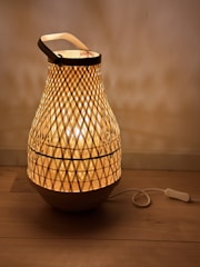 A lattice-patterned lamp with a warm glow, made from woven material. It has a rounded shape, resembling a basket or lantern, and sits on a wooden surface. A power cord with a switch is visible on the floor next to it.
