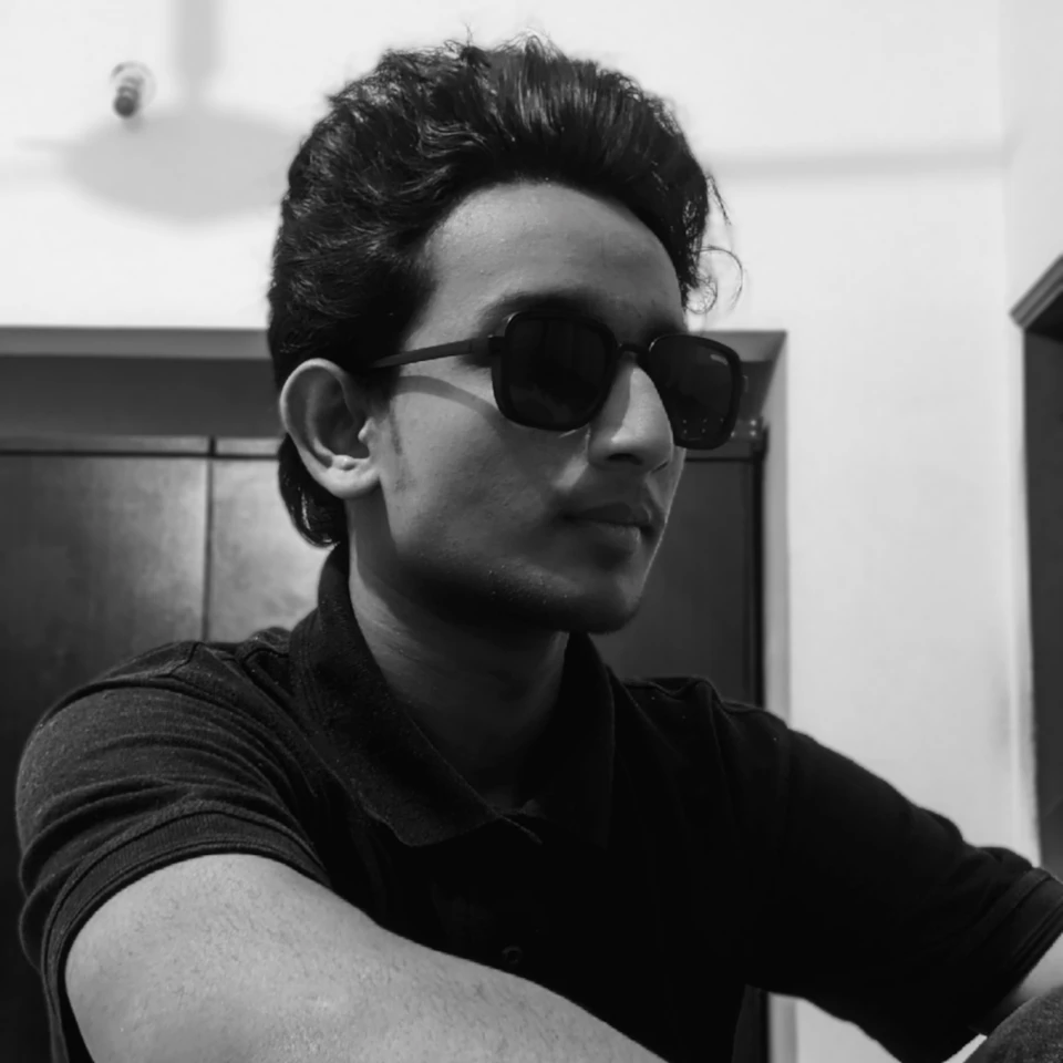Young man sits against white wall, wearing sunglasses and black polo shirt, gazing offside