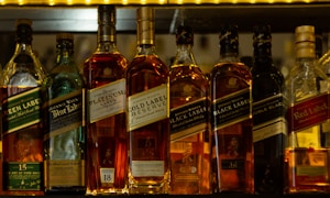 A selection of various labeled whisky bottles arranged side by side on a shelf, featuring different types such as Green Label, Blue Label, Gold Label Reserve, Black Label, Double Black, and Red Label. The scene is warmly lit with emphasis on the amber liquid inside the bottles.