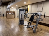 A well-lit gym with wooden flooring, showcasing a range of exercise equipment including weight machines. The room features a neutral color scheme with a focus on functionality and cleanliness. A sign with gym rules is mounted on the wall, and there's a counter with towels and disinfectant.