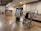 A well-lit gym with wooden flooring, showcasing a range of exercise equipment including weight machines. The room features a neutral color scheme with a focus on functionality and cleanliness. A sign with gym rules is mounted on the wall, and there's a counter with towels and disinfectant.
