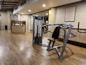 A well-lit gym with wooden flooring, showcasing a range of exercise equipment including weight machines. The room features a neutral color scheme with a focus on functionality and cleanliness. A sign with gym rules is mounted on the wall, and there's a counter with towels and disinfectant.