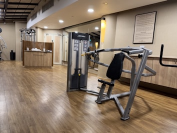 A well-lit gym with wooden flooring, showcasing a range of exercise equipment including weight machines. The room features a neutral color scheme with a focus on functionality and cleanliness. A sign with gym rules is mounted on the wall, and there's a counter with towels and disinfectant.