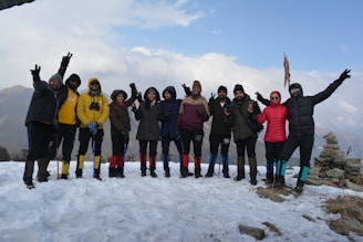 A group of people is standing on a snowy mountain, wearing winter clothing and hiking gear. They are posing together, with many showing peace signs, and appear cheerful and friendly. The sky is slightly cloudy, and there are distant mountains in the background.