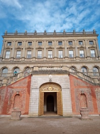 A grand and ornate building with classical architecture featuring large windows, detailed cornices, and decorative stonework. The entrance is highlighted by a golden gate, marked by intricate designs. The forefront shows an arched, symmetrically designed brick wall with planters on either side, and a gravel pathway leading to the entry.