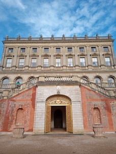 A grand and ornate building with classical architecture featuring large windows, detailed cornices, and decorative stonework. The entrance is highlighted by a golden gate, marked by intricate designs. The forefront shows an arched, symmetrically designed brick wall with planters on either side, and a gravel pathway leading to the entry.