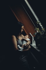 A woman is gently cradling a baby in her arms near a window with soft natural light. The setting is warm and intimate, with shadows accentuating the peaceful scene. Nearby, there is a potted plant adding a touch of nature to the cozy environment.
