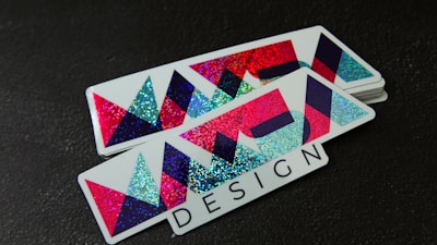 A stack of rectangular stickers featuring a geometric design with vibrant colors. The design includes overlapping triangles and other shapes with a glittery texture. The word 'DESIGN' is printed in a bold font at the bottom.
