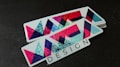A stack of rectangular stickers featuring a geometric design with vibrant colors. The design includes overlapping triangles and other shapes with a glittery texture. The word 'DESIGN' is printed in a bold font at the bottom.