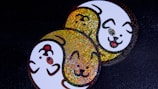 A pair of round stickers featuring cartoon dog faces intertwined in a yin-yang style. One dog face is in white with a smiling expression and a protruding tongue, outlined with glittery gold highlights. The other is primarily glittery gold with similar facial features. Both are set against a dark background, enhancing the vibrant colors.