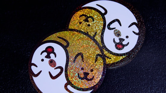 A pair of round stickers featuring cartoon dog faces intertwined in a yin-yang style. One dog face is in white with a smiling expression and a protruding tongue, outlined with glittery gold highlights. The other is primarily glittery gold with similar facial features. Both are set against a dark background, enhancing the vibrant colors.