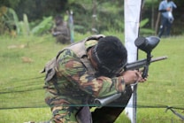 A person dressed in camouflage gear is crouched, holding a paintball gun, ready to shoot. They appear to be participating in a paintball game on a grassy field. Another person is visible in the background, partially out of focus, also participating in the game.
