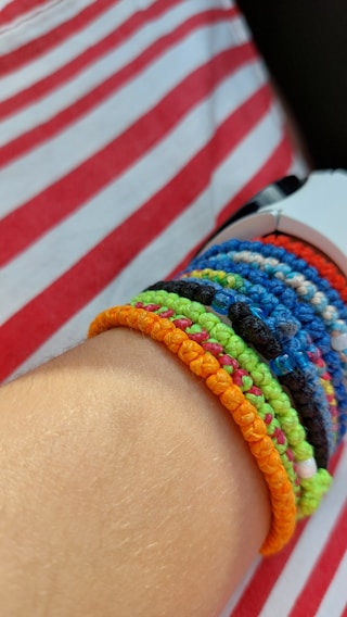 A forearm with multiple colorful woven bracelets made of different materials and colors, including orange, green, blue, black, and pink, against a background of a red and white striped shirt.