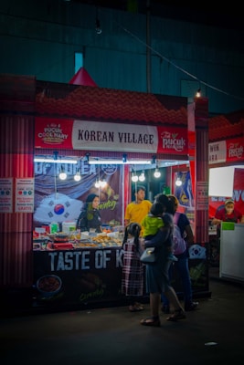 A food stall with a 'Korean Village' sign, decorated with elements related to Korean cuisine. The stall has a variety of food items on display, with a vendor behind the counter and customers, including a parent holding a child, observing the offerings. Bright lights illuminate the stall, creating a vibrant nightlife atmosphere.