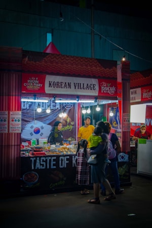 A food stall with a 'Korean Village' sign, decorated with elements related to Korean cuisine. The stall has a variety of food items on display, with a vendor behind the counter and customers, including a parent holding a child, observing the offerings. Bright lights illuminate the stall, creating a vibrant nightlife atmosphere.
