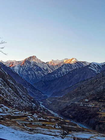 Snow-capped peaks glowing golden at sunset above a quiet valley dotted with traditional Himachali houses.
