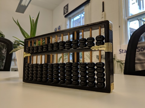 A traditional wooden abacus with black beads sits on a white tabletop in a well-lit room. In the background, there are potted plants and a window showing an outside view. Office furniture such as chairs and a desk lamp are visible.