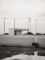 A parking lot is shown with a reflective puddle on the ground, surrounded by white concrete barriers. Several streetlights and power lines intersect the overcast sky. In the background, a small building structure with a flat roof is visible, while trees and a billboard can be seen in the distance.