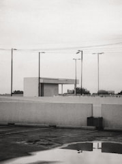 A parking lot is shown with a reflective puddle on the ground, surrounded by white concrete barriers. Several streetlights and power lines intersect the overcast sky. In the background, a small building structure with a flat roof is visible, while trees and a billboard can be seen in the distance.